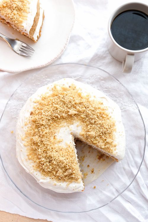 Top down view of a whole carrot cake with one slice removed, a slice of carrot cake on a plate, and cup of coffee on a table with a white tablecloth.