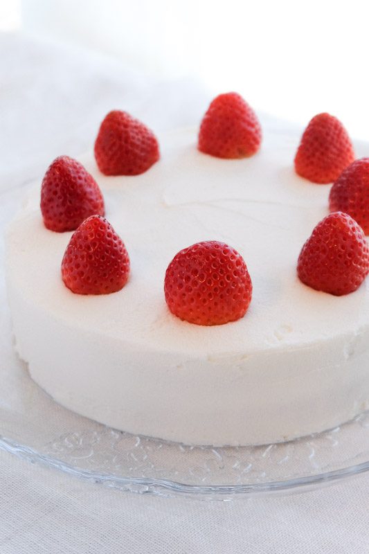 Side view of a vegan cake with white frosting and strawberries on a glass plate on a table with a white tablecloth. 