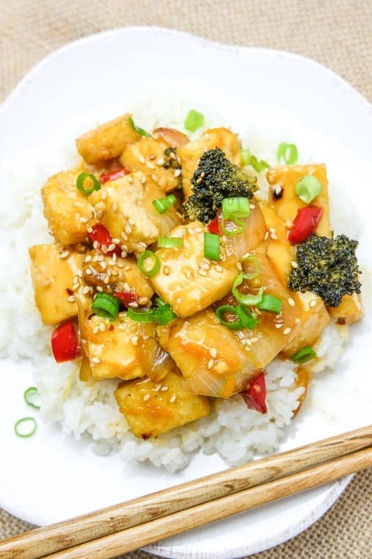 orange tofu with red peppers, broccoli, and green onion on a plate with rice and chopsticks.