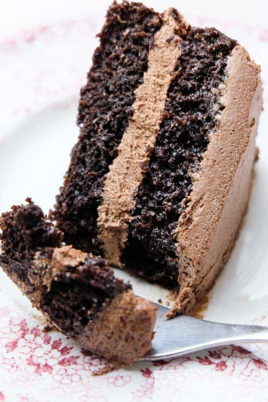 A vegan chocolate cake with chocolate frosting made from whipped coconut oil. A fork has taken a piece of the fluffy chocolate cake out and looks delicious.