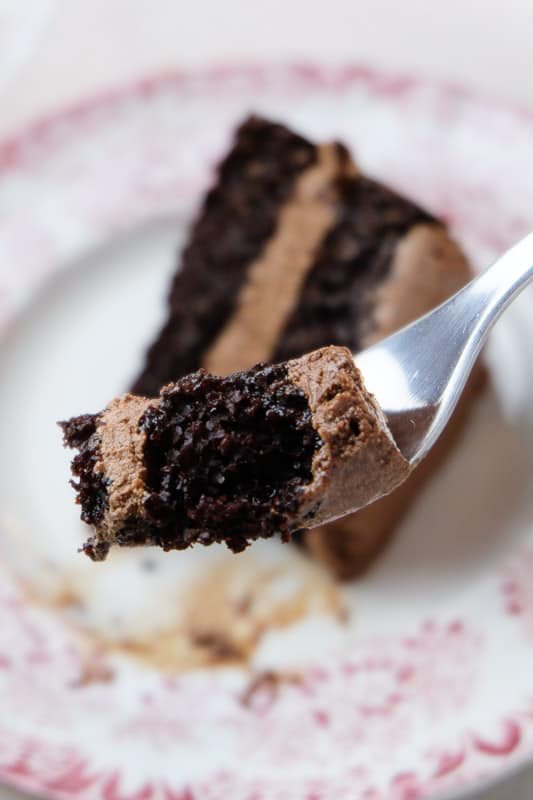 Close up shot of a bite of chocolate cake on a fork, with the rest of the slice of cake on a plate in the background.