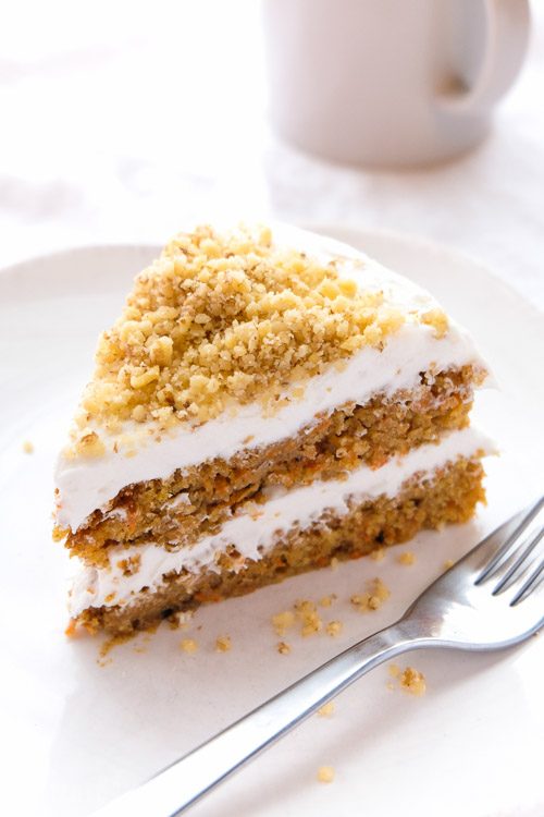 A slice of carrot cake with white frosting and chopped nuts on top, on a plate with a fork and a cup of coffee.