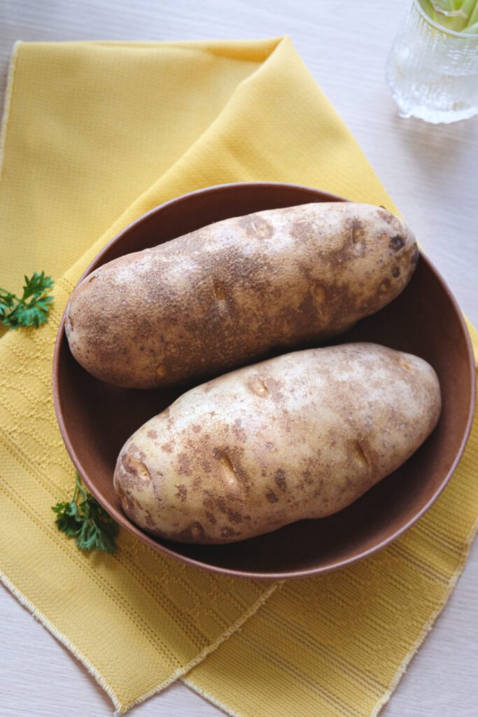 Two large russet potatoes in a bowl on a table with some parsley sprigs and green onions.