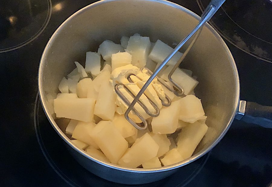 A potato masher pressing into a pot of cooked potato pieces, with a large pat of vegan butter.